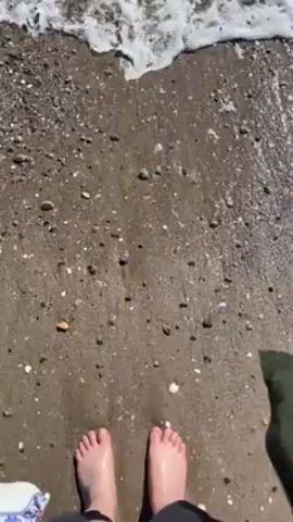 The video shows a first-person perspective from someone standing on a beach. The individual is barefoot and wearing what appears to be a dark-colored pair of shorts. Ocean waves wash over their feet and the sand. To the right, they carry a green object and to the left a white and blue patterned cloth.