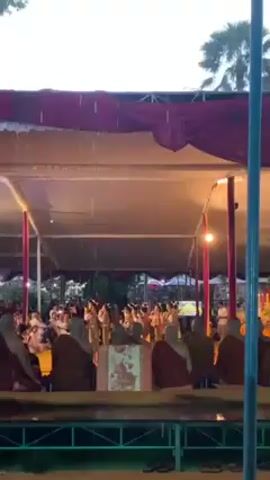 Here is a concise description of the video:

The video shows a group of women dressed in traditional attire dancing under a tent during a light rain. Seated in front of the dancers are several people in saffron robes, who appear to be observing the performance. The tent has a red and white roof and is lit by electric lights. In the background, trees and other structures are visible.