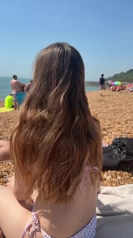 A young woman wearing a white polka dot bikini is sitting on a towel at a sunny beach. She moves her long brown hair. Other people are scattered across the pebble beach near the water. A black bag sits beside her.