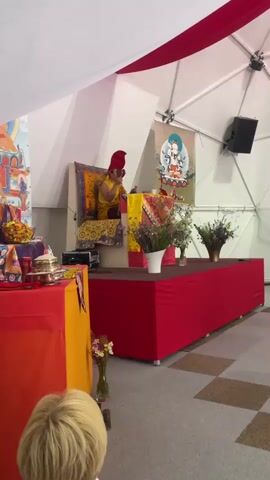 The video shows a Buddhist ceremony in progress. A person wearing traditional yellow robes and a tall red hat is seated on an elevated, ornate chair, playing a small, brown instrument. Another person, dressed in a maroon robe, stands nearby, holding a bundle of incense sticks. Both figures are positioned on a platform covered with a red cloth and surrounded by flowers. Behind the seated individual, there's a Buddhist tapestry and an image of a deity. The setting appears to be indoors, with the space decorated with white and red fabric. The room has a unique architectural design, possibly a dome structure. Sounds of percussion instruments accompany the actions of the individuals.