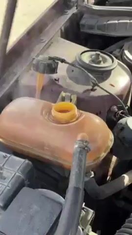 A person removes the coolant reservoir cap on a car. The reservoir has the words “KALT/COLD” inscribed on it. The person uses a dipstick-like instrument to check the coolant level, then replaces the reservoir cap. The car’s hood is open, showing the engine.