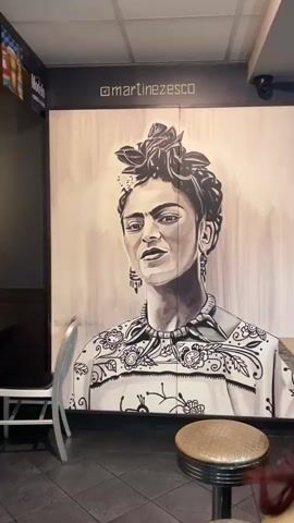 The video showcases a large, black and white mural of Frida Kahlo in an indoor setting, likely a restaurant or bar. The mural depicts Frida Kahlo with her signature unibrow, braided hair adorned with leaves or flowers, distinctive earrings, a necklace, and a patterned top. Her expression is serious and direct.

The video begins with a wide shot of the mural, showing a bar stool with a chrome base and a textured, possibly leather, seat in the foreground, and a silver-colored chair to the left. Above the mural, the text "@martinez_esco" is visible, likely an artist's or establishment's social media handle. A partial "Modelo" advertisement can be seen on the left wall.

The camera then zooms in on Frida Kahlo's face, highlighting the details of the artwork. Following this, there's a brief, blurry shot of colorful lights or reflections. The video concludes with a close-up of a speckled, light-colored surface, possibly a countertop or table, showing its texture and a subtle reflection. The indoor lighting suggests the establishment is open, but the specific time of day is not discernible.