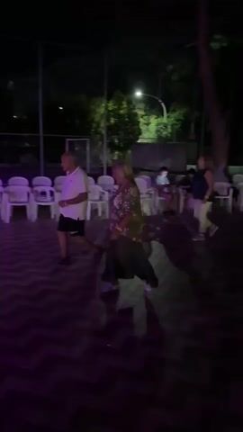 The video shows an outdoor nighttime scene. In the foreground, a patterned tiled dance floor is illuminated by purple and white lights. Several people are visible dancing or standing on the dance floor. A man in a white polo shirt and dark shorts is dancing. A woman with blonde hair, wearing a patterned top and a long green skirt, is also dancing. Another woman in a dark top and light pants stands further back. In the background, white plastic chairs are arranged around tables. A wire fence, likely for a sports court, is visible behind the chairs. Trees and foliage are present in the background, and a streetlight provides illumination.