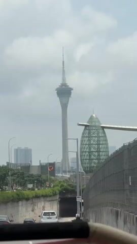 The video shows a view from a moving vehicle towards the Macau Tower, a tall communication and observation tower in Macau, China. In the foreground, there is a large, green, egg-shaped structure. There is a tunnel underneath the road, with cars entering and exiting. The sky is overcast. A white car drives through the tunnel.