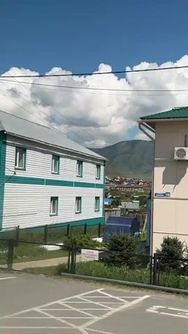 The video shows a moving shot from a vehicle, possibly a car, looking out of a window. The camera pans across a village or small town setting. The first building visible is a light-colored, two-story building with a green roof. It has several windows and air conditioning units on its exterior. A blue and white sign with a "P" and a wheelchair symbol is visible on the left, along with a sign indicating a 2-meter width limit. Further down the street, another building with a green roof and multiple windows is seen, with a white and green house with a light-colored roof next to it. The camera then shows a wooden fence and a glimpse of houses with red roofs and mountains in the background. Road signs are visible, including a red circular sign with a white horizontal bar and a yellow triangular sign with black dots. Trees and foliage are present throughout the scene. The sky is blue with scattered white clouds.