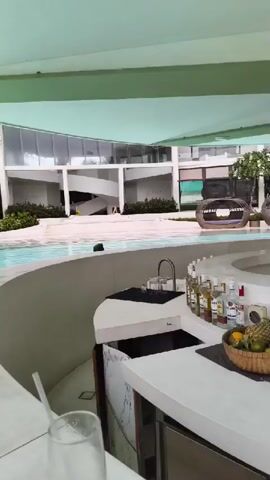 A high-angle, medium shot shows a resort-style pool bar area. The bar itself, made of white, marble-patterned material, is in the foreground. On the counter, a basket of fruit sits next to an assortment of liquor bottles. Behind the bar, a sink and various bar tools are visible. A swimming pool curves around the bar area, with lounge chairs in the background. A building with modern architecture, featuring large windows and a spiral staircase, is visible beyond the pool. The sky is overcast, with a light green awning extending over the bar. A person is briefly seen swimming in the pool.