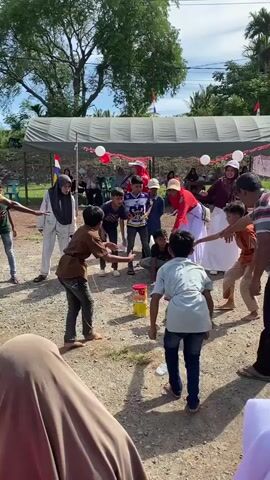 A group of children, mostly boys, are participating in a game outdoors. They are standing on a dirt ground, with a tent and decorations in the background, suggesting a festive occasion. In the center of the activity, there are two stacks of colorful cans, possibly for a game of knocking them down. Some children are holding ropes or strings, which might be part of the game's mechanics, like pulling or throwing. Several adults are observing the game, some standing and some sitting under the tent. The atmosphere appears to be lighthearted and engaged.