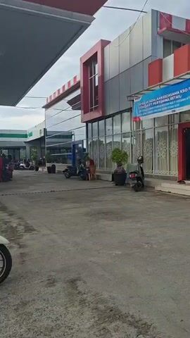 The video pans across the exterior of a building. A person wearing an orange shirt and blue jeans is seated on a white motorcycle. They are wearing a white helmet and appear to be looking to their left. The motorcycle is parked on a paved surface. In the background, there is a building with large glass windows and a red and white facade. Several other motorcycles are parked in the distance. There is also a large banner hanging on the front of the building. The overall environment appears to be an outdoor commercial area.