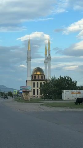 The video captures a mosque with a golden dome and four tall white minarets with golden spires. The mosque is a light-colored stone building with several windows. To the left of the mosque, there is a road with cars driving by and some billboards. A bush and a white shipping container are visible to the right of the mosque. In the foreground, several cows are walking on the side of the road. The sky is partly cloudy with a mix of blue sky and white clouds. There are also some hills in the background. The overall scene appears to be a rural or semi-rural area, possibly in the evening given the lighting.