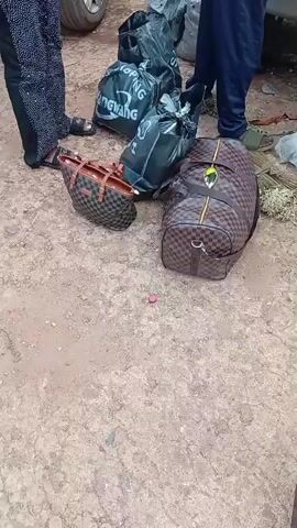 The video shows a group of people loading luggage into and around two silver cars parked on a dirt road. In the foreground, there are several large bags, including a brown duffel bag with a checkered pattern and a yellow crest, and a large white bag labeled "GROWER FEED". Behind these, multiple black trash bags are visible. Two individuals, a man wearing a dark blue tracksuit and a woman in a blue t-shirt and patterned skirt, are near the back of the first car. The man appears to be putting a large, clear plastic bag filled with items into the open trunk. A second car is visible behind the first, with its trunk also open and a person in the process of loading bags into it. A woman in a patterned headscarf and colorful skirt stands near the second car, observing. The environment appears to be outdoors, possibly in a rural area, with vegetation and buildings visible in the background. The sky is overcast.