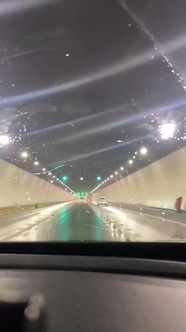 The video shows a point-of-view shot from inside a vehicle driving through a tunnel. The tunnel is illuminated by numerous lights along the ceiling and walls. Green directional arrows are visible above the lanes, indicating the flow of traffic. Several cars are driving in the same direction. The road surface appears wet, suggesting it may be raining or has recently rained. The windshield has some visible streaks or marks, which could be water droplets or dirt. The overall environment is dark and enclosed, characteristic of a tunnel.