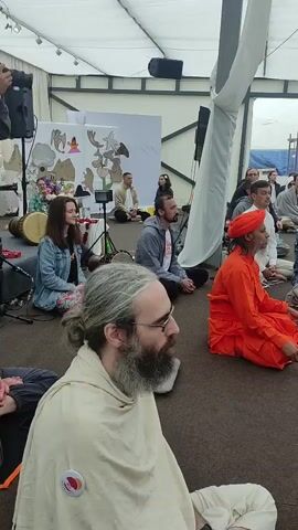 The video shows a gathering of people sitting on the floor in what appears to be an open-air pavilion. The participants are mostly seated in a cross-legged meditation posture, facing a central point where a small altar or display is visible. The environment suggests a retreat or spiritual event. The people are dressed in casual to semi-formal attire, with some individuals wearing orange robes, possibly indicating a specific spiritual tradition. The overall atmosphere seems calm and focused.