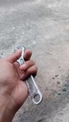 The video shows a hand holding an adjustable wrench. The hand is seen from a close-up perspective, and the wrench is manipulated to show different angles. The background appears to be a concrete surface. The wrench is made of metal and has a silver finish. It is an adjustable wrench, which means its jaw size can be changed to fit different nuts and bolts. The hand is wearing a black ring on one of its fingers. The video is brief, lasting only a few seconds.