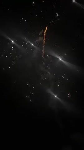 The video shows fireworks exploding in the night sky. A rocket shoots up, leaving a trail of sparks, and explodes into a starburst of light. There are sounds of explosions and crackling. The sky is dark with some light in the distance.