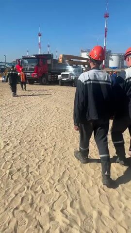 Two construction workers in hard hats and reflective vests walk across a sandy area. In the background, there is a red truck, a crane, and several large industrial tanks under a clear blue sky. The ground is covered in sand with visible footprints and tire tracks. The workers appear to be walking towards the truck and other equipment.
