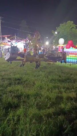 The video shows a small, old-fashioned carousel operating on a grassy field at night, with other amusement park rides and tents visible in the background, illuminated by streetlights and ride lighting. The carousel features animal figures, including horses and a camel, and is spinning but not occupied by riders. The camera angle is from ground level, slightly off-center, and captures the movement of the carousel against the backdrop of the night fair.