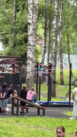 A person is performing a gymnastic routine on a bar in an outdoor park. The person, wearing a teal shirt and grey pants, is in mid-air, performing a flip. They are surrounded by trees, with a group of spectators watching from a bench. There are other pieces of exercise equipment visible in the background.