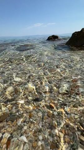 This is a vertical, first-person perspective video of a rocky beach and clear, shallow ocean water. The scene is bathed in bright sunlight, suggesting it's daytime, likely midday given the clear, vibrant blue sky with only a few wisps of clouds. The water is so transparent that the colorful pebbles and stones on the seabed are clearly visible, shimmering and shifting with the gentle movement of the waves. Two dark, weathered rocks rise out of the water in the middle ground. In the distance, a hazy, landmass can be seen on the horizon, with the expanse of the ocean stretching out to meet the sky. The overall impression is one of tranquility and natural beauty, a serene coastal environment. No people, objects, or text are visible in the video.