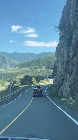 A vehicle is driving on a winding mountain road. The road is paved and has yellow and white lines. A large rock face is on the right side of the road. Mountains covered in trees are visible in the distance on the left. The sky is blue with some clouds. Another car is visible ahead, also driving on the road.