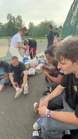 The video depicts a group of young adults sitting on asphalt in an outdoor setting, possibly a skate park, with a woman distributing items from a cooler. There are at least seven people visible, a mix of genders and ethnicities, mostly dressed in casual streetwear. Food items and a drink can are scattered around them. A laptop placed on the ground is playing a soccer game on its screen, showing a close-up of a player in an Arsenal jersey who has just scored, followed by a crowd scene. Graffiti is visible on a nearby structure.