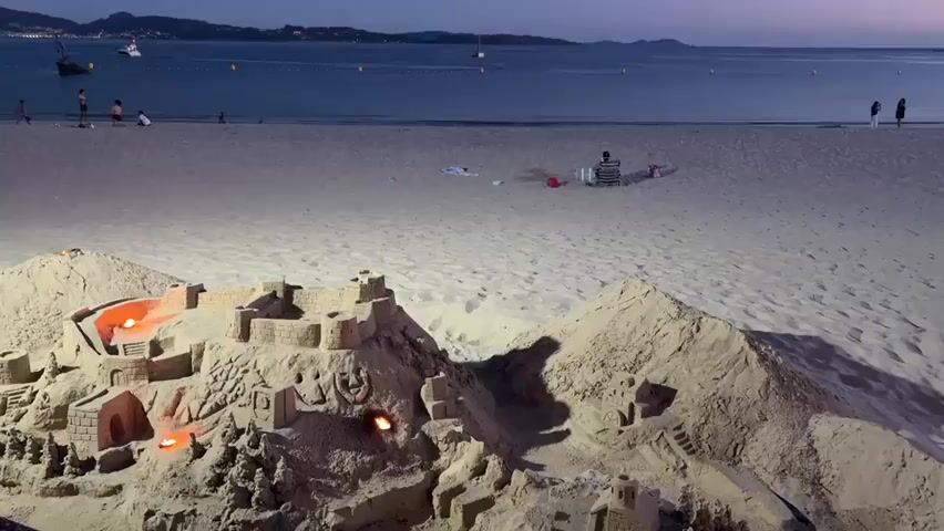 The video shows a sandy beach at dusk. In the foreground, a large, elaborate sandcastle complex dominates the frame. The sandcastle has multiple towers, walls, and intricate details, including what appear to be carved faces and small doorways. Small fires are burning within some of the structures, casting an orange glow. To the right of the main sandcastle, there is a large, conical sand dune, and further to the right, a smaller sand structure resembling a building. The beach is a pale sand, and in the middle ground, there are a few scattered beach items like towels and chairs. Further back, a calm body of water stretches to the horizon. A few small boats are visible on the water, and a distant coastline with hills is dimly lit against the twilight sky. Several silhouetted figures of people can be seen walking on the beach in the distance, and a few children are playing closer to the water's edge. The overall ambiance is peaceful and serene, with the twilight lighting and the gentle glow of the fires creating a slightly magical atmosphere.
