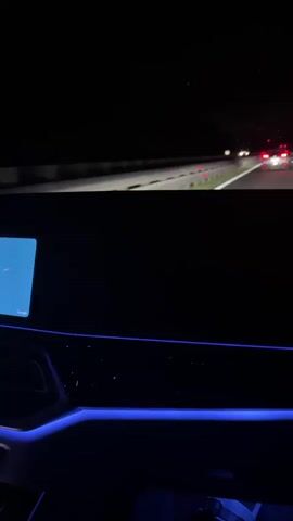 The video shows a first-person view from inside a car at night, driving on a highway. The top half of the screen shows the road ahead, with other cars visible in the distance, their taillights illuminated. The bottom half of the screen shows the car's interior, which is dimly lit and features blue ambient lighting. A screen, possibly a navigation display, is visible on the left side of the dashboard. The overall impression is of a night drive on a highway.