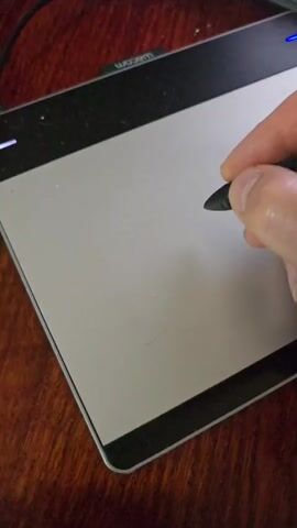 A hand holds a stylus and draws on a Wacom drawing tablet. The tablet is silver with a black border and the Wacom logo is visible at the top. The drawing tablet rests on a wooden surface.