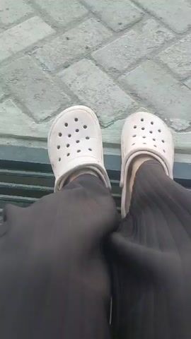 This is a first-person, overhead, static shot of a person's feet wearing light-colored Croc-style shoes and dark, possibly black, pants. The shoes are resting on a surface made of light gray rectangular paving stones arranged in a herringbone pattern. A dark metal grate is visible beneath the paving stones. The person's feet are slightly angled, with the left foot positioned slightly in front of the right. The lighting suggests it is daytime. The video appears to be a short, still recording with no discernible action.