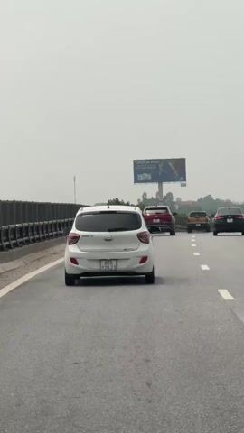 The video shows a white Hyundai car driving on a highway, as seen from the driver's perspective. The car is in the slow lane, following a line of other vehicles including a red SUV, an orange SUV, and a black sedan. On the left is a metal safety barrier. On the right of the highway are roadside billboards, displaying advertisements in Vietnamese. The sky is overcast. The road is paved and marked with white lines separating the lanes. The car's license plate is visible.