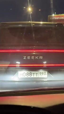 A vehicle's perspective from inside a car, focused on the tail end of a dark-colored Zeekr car moving on a highway at night. The Zeekr's taillights are illuminated, and the license plate is visible. Streetlights illuminate the highway. On the right side of the frame, a billboard is partially visible. Other vehicles are present on the highway. The video is taken through the windshield of the car.