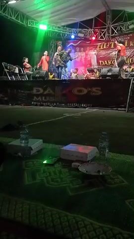 The video shows an outdoor stage performance at night. A band or musical group is on stage, with several people singing and playing instruments. There are lights illuminating the stage and a banner behind the performers. In the foreground, there are several water bottles and boxes on a mat. The overall atmosphere appears to be a lively event.
