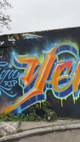 The video shows a long wall covered in graffiti artwork. The wall is mostly dark gray or black, but some sections are pink or white. The graffiti is colorful, with lettering in orange, blue, green, purple, and yellow. Some of the graffiti includes tags such as "217 S.V.P.P" and "LSC Savor Latino." There is grass and weeds growing at the base of the wall and some concrete structures in front of the wall. The sky is overcast. The camera pans along the wall to show the different graffiti pieces.