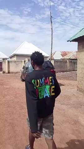 A person wearing a black long-sleeved shirt with "SUMMER PEACE" in multicolored letters, camouflage shorts, and sandals walks away from the camera on an unpaved, dusty road. They are holding a dark object in their right hand, swinging it as they walk. In the background, a dark green SUV is parked. Several houses with white, green, and reddish-brown roofs are visible. A utility pole with wires stands to the left. Near the end of the clip, another person in a blue and white patterned garment is partially visible on the right side of the frame near a building. The sky is partly cloudy.