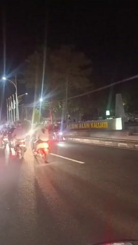 The video is a nighttime scene of a street. A dark-colored pickup truck with a cage-like structure in the back is moving on the right side of the road. Its taillights are illuminated. Several motorcycles are traveling in the same direction on the left side of the road. Streetlights and other artificial lights illuminate the scene, creating lens flares and reflections on the wet road surface. Trees are visible in the background. There are some signs in the background, one of which appears to have text in Indonesian.