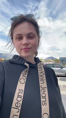 A woman stands outside in windy conditions, smiling. She wears a dark Calvin Klein Jeans hoodie and has her hair pulled back. A McDonald's restaurant is visible in the background, along with mountains. A silver car is parked in the background. The sky is cloudy.