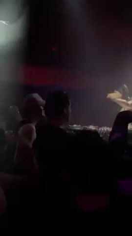 The video shows a female DJ performing at a nightclub. She wears a bikini top and stands behind a DJ console, adjusting her headphones and working the equipment. The club is dark, with red accent lighting, and smoke fills the air. A crowd of people faces the DJ, their backs mostly to the camera. Some are moving and raising their hands in enjoyment.