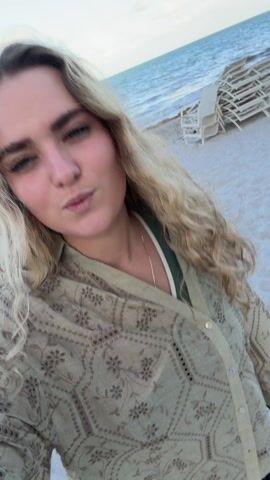 A woman with blond curly hair wearing a tan patterned shirt and a black skirt is filming a selfie video on a sandy beach. In the background, a gray trash can is visible. The sky is blue. The woman smiles at the camera and gives a kissy face.