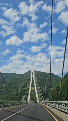 A first-person view from a vehicle on a road approaching a modern cable-stayed bridge. The sky is blue with scattered white clouds. The bridge has two tall white support towers, from which numerous cables descend to the roadway. The roadway is asphalt with yellow and white lane markings. Mountains covered in green trees rise behind the bridge. Streetlights are visible on the bridge. There are no people visible, but a dark-colored vehicle is seen on the bridge in the distance. The camera is moving forward.