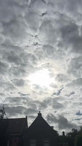A vertical shot of a sky filled with dramatic, textured clouds. The sun shines brightly through a break in the clouds, creating a halo effect. In the foreground, the silhouette of a dark-colored house with a gabled roof is visible. To the left, a portion of another house with a dark roof and solar panels can be seen. A single bird is flying in the upper portion of the sky. The overall impression is a cloudy day with the sun attempting to break through.