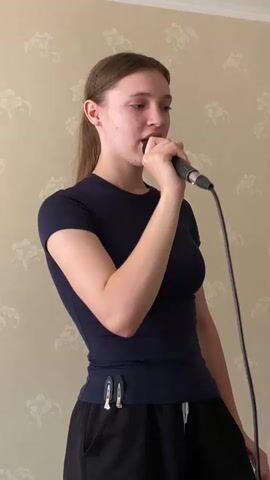 A young Caucasian female with light brown hair pulled back in a ponytail is standing and singing into a microphone. She is wearing a dark blue short-sleeved shirt and black shorts. The background is a wall with a light-colored floral wallpaper pattern. She is holding the microphone with her right hand near her mouth. There are two white clips attached to the waistband of her shorts. The video is shot from a medium distance, showing her from the waist up.