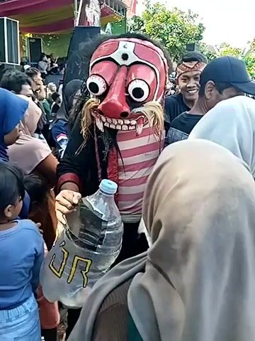 The video shows a person wearing a large, grotesque red mask with exaggerated features such as bulging eyes, a bulbous nose, and a wide, toothy grin, surrounded by a crowd of people. The masked figure is wearing a horizontally striped pink and white shirt over a black garment. They hold a large plastic water bottle with writing on it. The crowd includes men, women, and children, some of whom are smiling or looking curiously at the masked individual. 

A stage with sound equipment and colorful decorations is visible in the background. The setting appears to be outdoors, possibly at a cultural event or festival. The lighting suggests it is daytime. The camera is handheld and moves slightly.