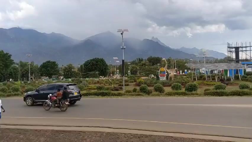 The video shows a scenic view of a roundabout with a statue of two giraffes, two human figures, and an emblem, situated amidst a landscape of green shrubbery and yellow flowers. In the background, a mountain range shrouded in misty clouds provides a striking backdrop. Overhead, a network of solar panels is visible. Vehicles, including cars and a three-wheeled auto-rickshaw carrying passengers, are seen navigating the road surrounding the roundabout. The overall atmosphere of the video is calm and picturesque.