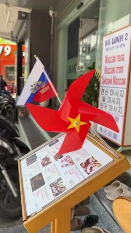 The video shows a close-up view of a red pinwheel with a yellow star in the center, mounted on a wooden stand displaying a menu. Behind the pinwheel, two flags are visible: one with the Russian flag and another with the Vietnamese flag. The menu displays images of people receiving massages. In the background, a building with signs in Vietnamese and possibly Russian can be seen, along with some parked motorcycles. The overall environment appears to be an outdoor area in front of a business, likely a spa or massage parlor. The pinwheel is spinning.