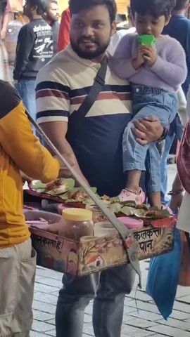 The video captures a lively street scene in Manali, India. The central focus is a man holding a young child. The man, with a beard and wearing a striped t-shirt, is looking towards the viewer with a slight smile. He is carrying the child, who is dressed in a lavender sweater and blue jeans, and appears to be eating something green from a small cup or cone.

To their left, a vendor is preparing food on a cart. The cart is adorned with colorful paintings and has various containers and items for sale, including what appears to be paan, a traditional Indian mouth freshener or digestive. Some of the items are placed on green banana leaves. The vendor, wearing a yellow and black shirt, is actively engaged in preparing the items.

The background shows a bustling crowd of people, suggesting a busy public area, possibly a marketplace or a tourist spot. The lighting suggests it might be daytime or early evening. The overall atmosphere appears casual and vibrant, typical of a street-side commerce setting in India.

Visible text on the cart includes "क. क. ताहे" and "क. क. पान" which translates to "K. K. Paan" or "K. K. Tale" and "K. K. Pan", indicating the name of the vendor or the shop. The general setting and the items being sold strongly suggest a food stall on a street in India, with the specific location identified as Manali.