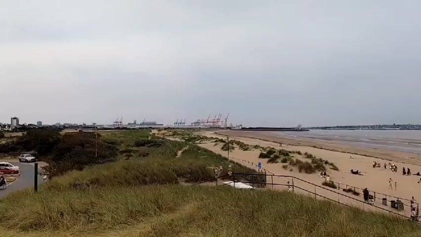 The video pans across a beach and a large, dome-shaped building. The sky is overcast. In the foreground, tall grasses are visible. To the left, a parking lot is filled with cars, and some buildings line the coast. A beach with several people walking and sitting on the sand stretches into the distance. Further out to sea, large cranes are visible, suggesting a port or industrial area. The dome-shaped building appears to be a modern structure with large windows and is surrounded by a parking area.
