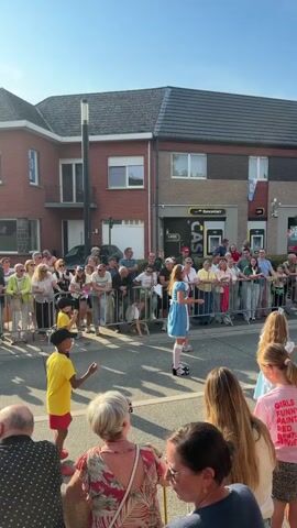 A group of people are gathered on a street, separated from a road by metal barriers. On the road, two children in bright yellow shirts and red shorts are dancing. In the background, a woman in a blue dress is standing and facing the children. She is also separated from the road by barriers and a crowd of onlookers. A building with a brick facade and a dark tiled roof is visible behind the crowd. The sky is clear and blue, and the sun is shining. The scene appears to be an outdoor event, possibly a festival or parade.