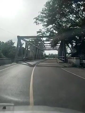 The video begins by showing a view from inside a vehicle traveling across a metal truss bridge. The bridge has a road surface and railings on either side. Palm trees and other foliage are visible on the banks of what appears to be a body of water under the bridge. As the vehicle continues, the bridge ends, and the road opens up into a more rural or semi-urban area with scattered buildings and more palm trees. Several motorcycles and a few other vehicles are seen on the road ahead and to the sides. The sky is overcast. The camera perspective remains fixed from the vehicle's interior, suggesting a dashcam or similar recording device.