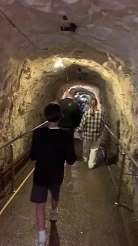 A group of people is walking through a long, narrow tunnel. The tunnel has rough, uneven walls, and a low ceiling. The floor is wet and there are metal railings on both sides. The group consists of a boy in a black jacket and shorts, a man in a black jacket, a woman in a plaid shirt and white pants, and others ahead of them. The lighting is dim, but there are some light fixtures on the ceiling. The group is walking in the same direction, towards the end of the tunnel.