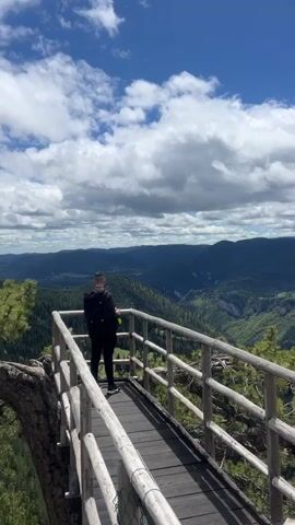 The video opens with a man standing on a wooden platform with a wooden railing, overlooking a vast mountain range under a partly cloudy sky. The man is wearing a black jacket and pants, and has a backpack on. He appears to be holding a green object in his hand. The camera slowly pans to the right, focusing on the panoramic view of the forested mountains. The clouds move across the blue sky, creating shadows on the landscape below. The wooden platform is secured with a wire mesh fence.