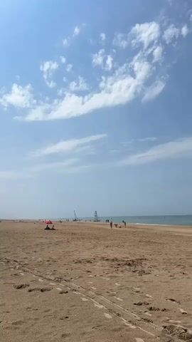 This media file captures a wide-angle view of a sandy beach on a bright, sunny day. The sky is a clear blue, dotted with wispy white clouds that stretch across the horizon. The beach is wide and appears to be relatively uncrowded, with the sand showing tracks from vehicles or ATVs.

In the distance, the calm sea meets the horizon, its surface reflecting the bright sky. Several people are visible on the beach, scattered across the sand. Some are walking along the shoreline, while others appear to be relaxing. A red umbrella provides shade for a person or group seated on the sand closer to the foreground. Further out in the water, a few sailboats and what appear to be windsurfing boards are visible, suggesting some water sports are taking place. The overall scene conveys a sense of peacefulness and leisure, typical of a summer day at the beach. The time of day is likely midday or early afternoon, given the bright sunlight and the angle of the shadows. The location is Koksijde, Belgium, a coastal town known for its beaches. There are no discernible texts within the frame.